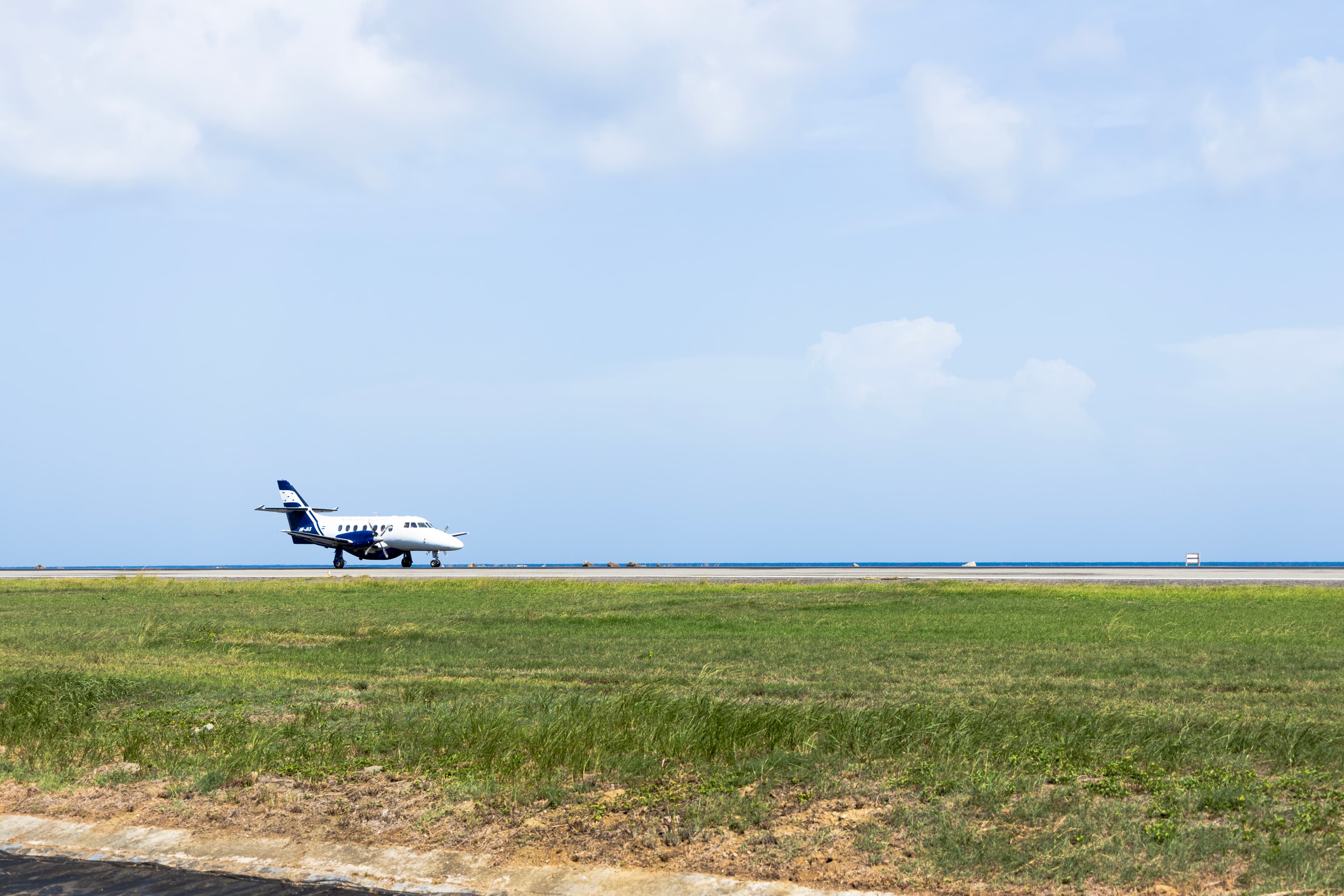 A small commercial airplane taxiing on a runway with a backdrop of a clear blue sky and ocean. The foreground features a grassy field, highlighting the airport's coastal location.