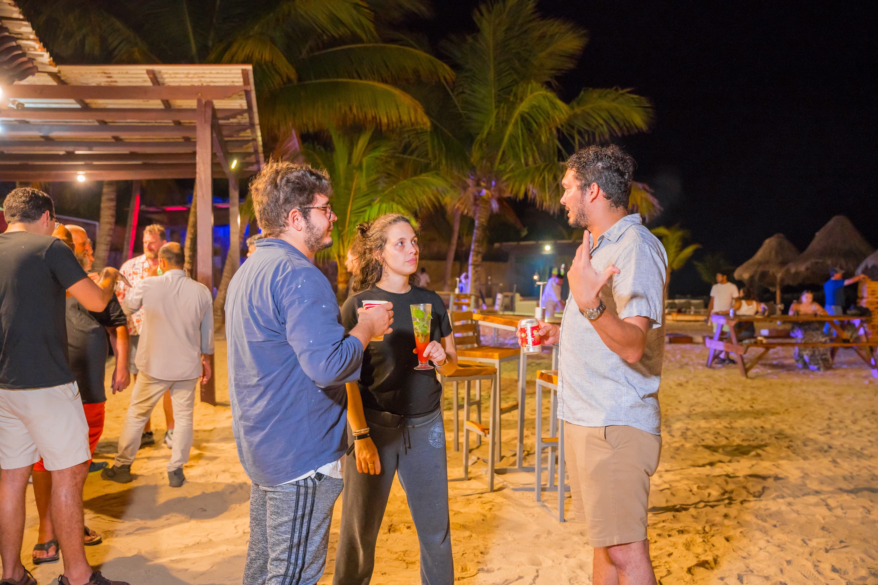 Group of people on a social gathering talking on a beach with drinks on their hands.