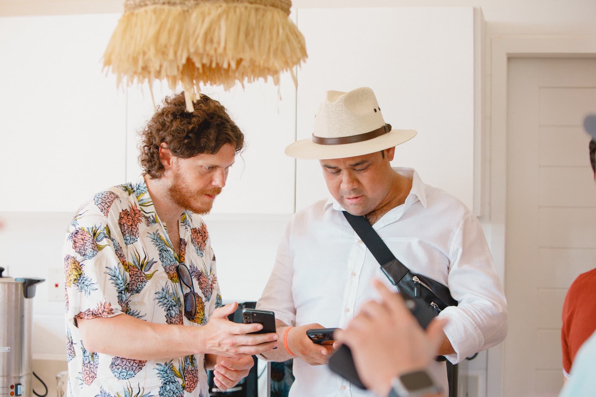 Two men standing indoors, closely looking at their smartphones, one wearing a straw hat and a crossbody bag, and the other in a floral shirt. They appear to be discussing or sharing something on their phones, creating an atmosphere of casual engagement.