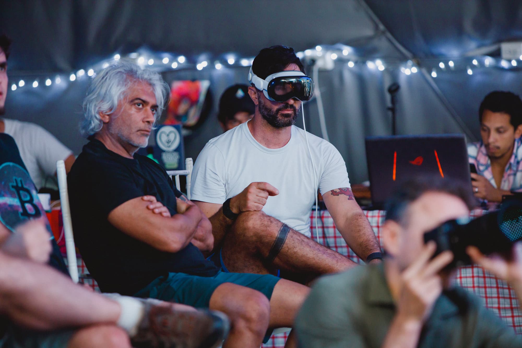 A man wearing a virtual reality headset sitting next to another man, both appearing focused and engaged. The setting is casual, with string lights in the background, indicating a relaxed and tech-oriented environment.