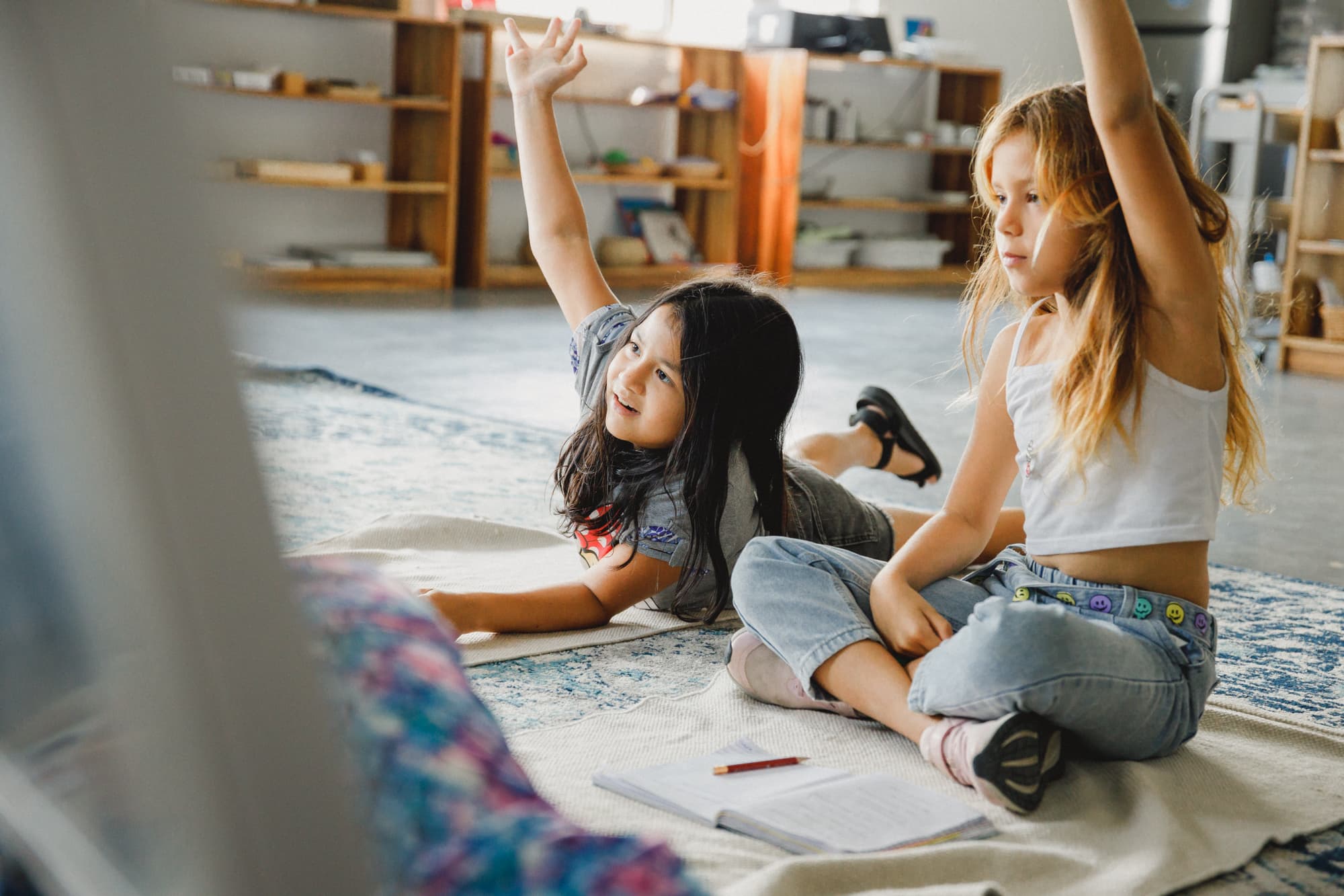 Two girls sitting on the floor in a classroom, raising their hands and engaging in a lesson. The environment is relaxed, with books and materials scattered around, and shelves filled with educational resources in the background.