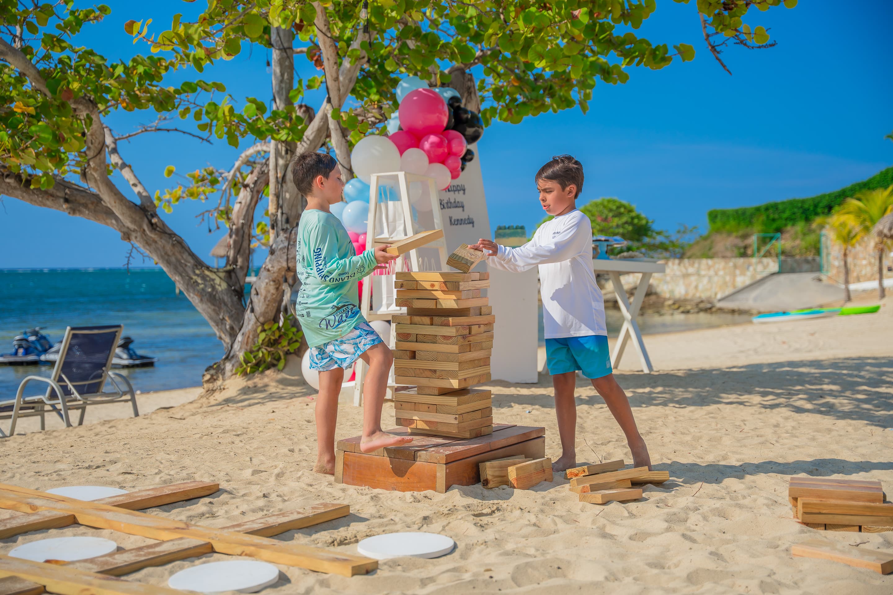 Two boys playing a game of giant Jenga on a sandy beach, with a backdrop of trees and balloons. The scene is bright and sunny, featuring a festive setup with colorful decorations and a beautiful ocean view in the background.