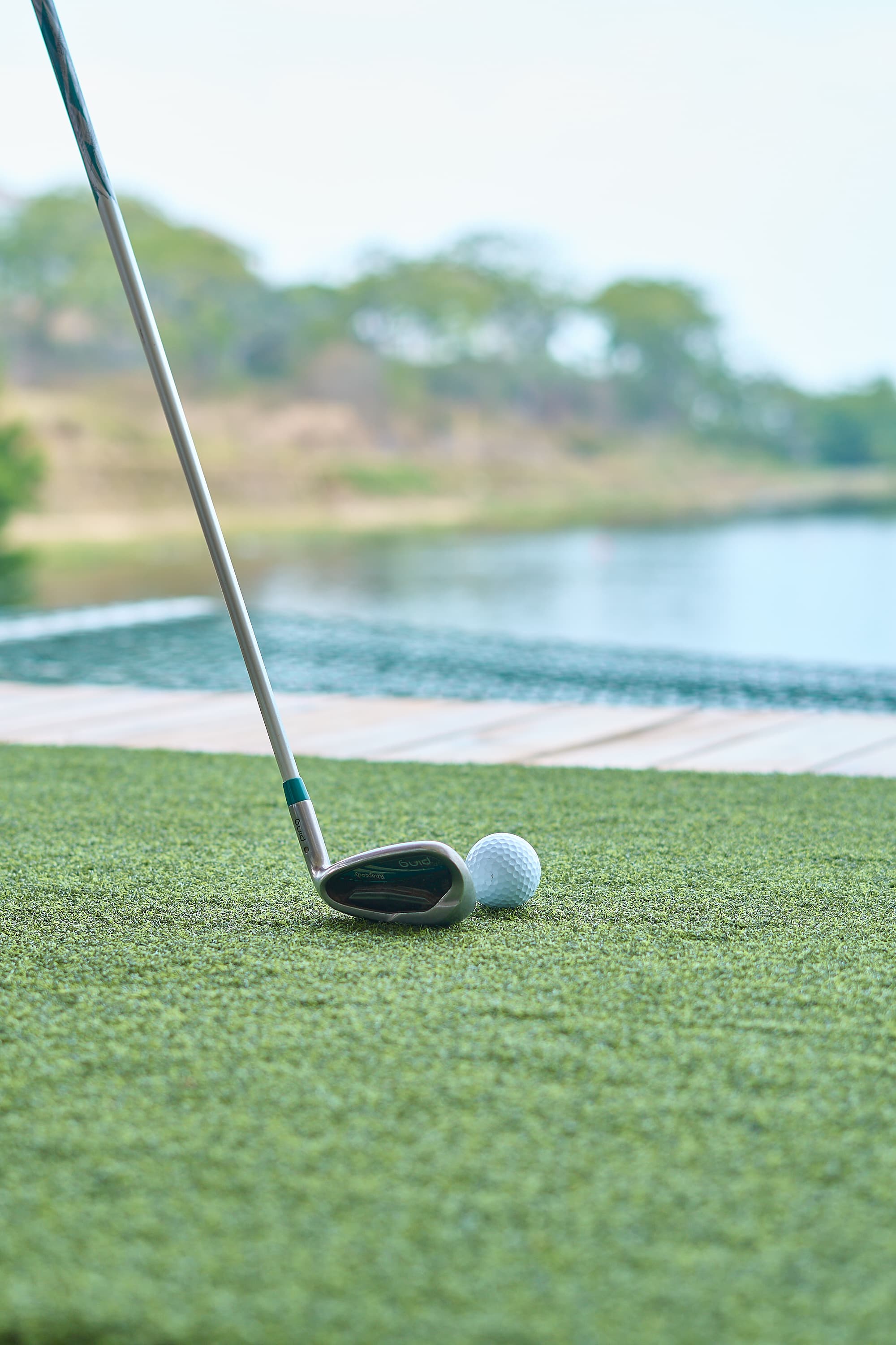 Close-up of a golf club and ball on an artificial turf near a water hazard on a golf course. The calm water and greenery in the background create a peaceful and focused setting for a game of golf.