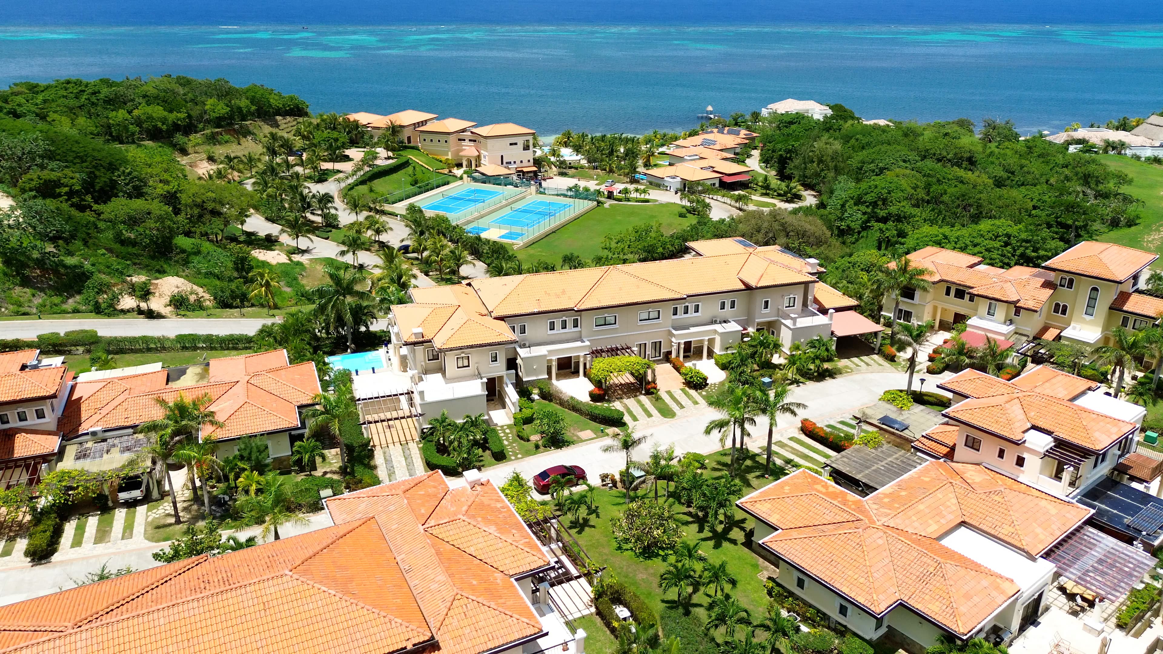 Aerial view of a coastal resort featuring multiple buildings with terracotta roofs, tennis courts, and lush green landscaping. The resort is situated next to a blue ocean, with the beach and a dock visible in the background, creating a picturesque and luxurious setting.