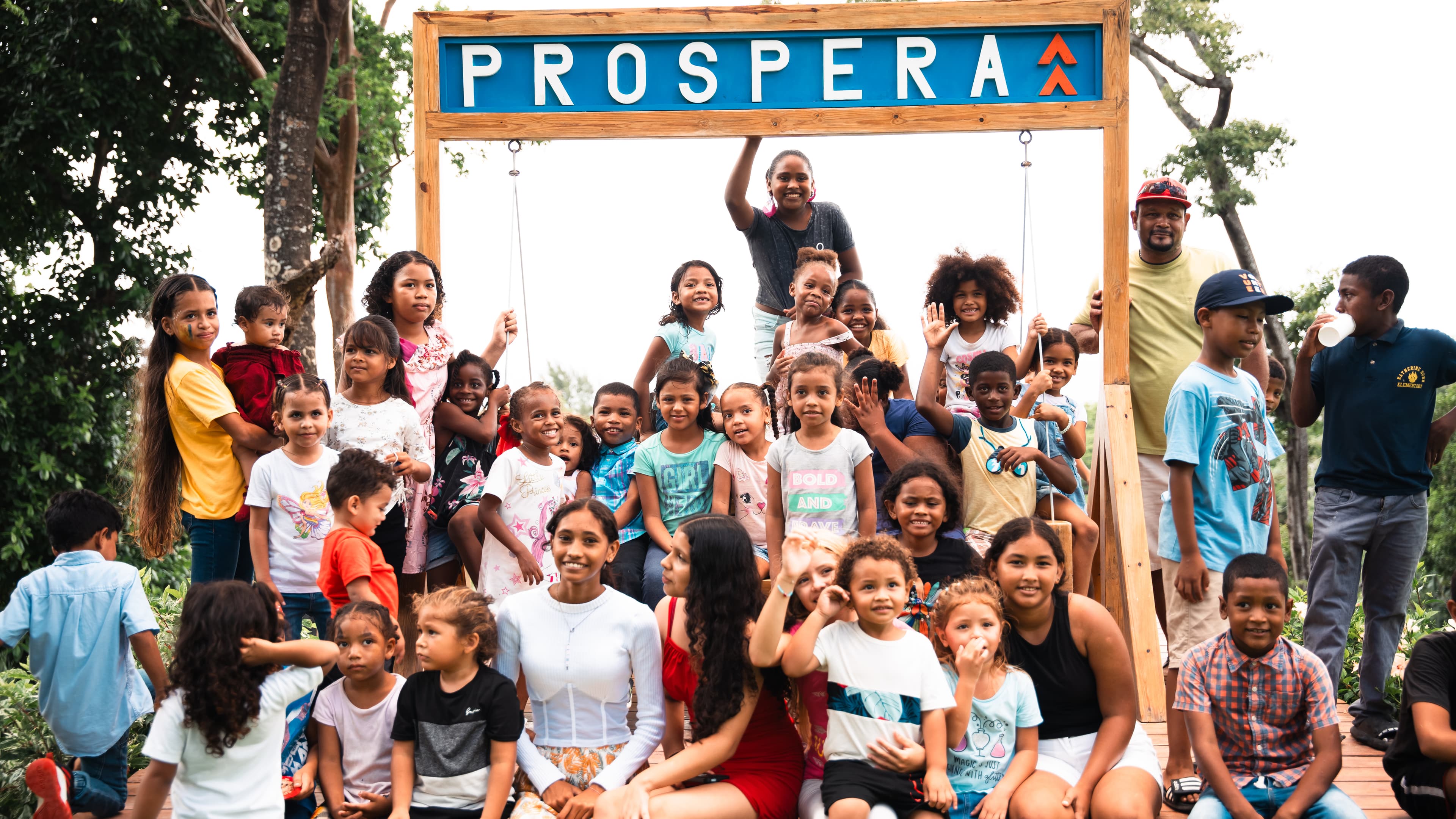 A large group of children and a few adults gathered under a wooden sign that reads PROSPERA, smiling and posing for the camera. The setting appears to be a community event or celebration, with everyone looking cheerful and engaged.